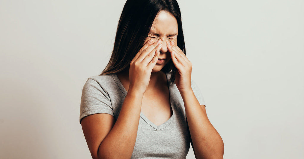 A long-haired woman in a gray short-sleeved shirt is rubbing her eyes while standing in front of a gray background.