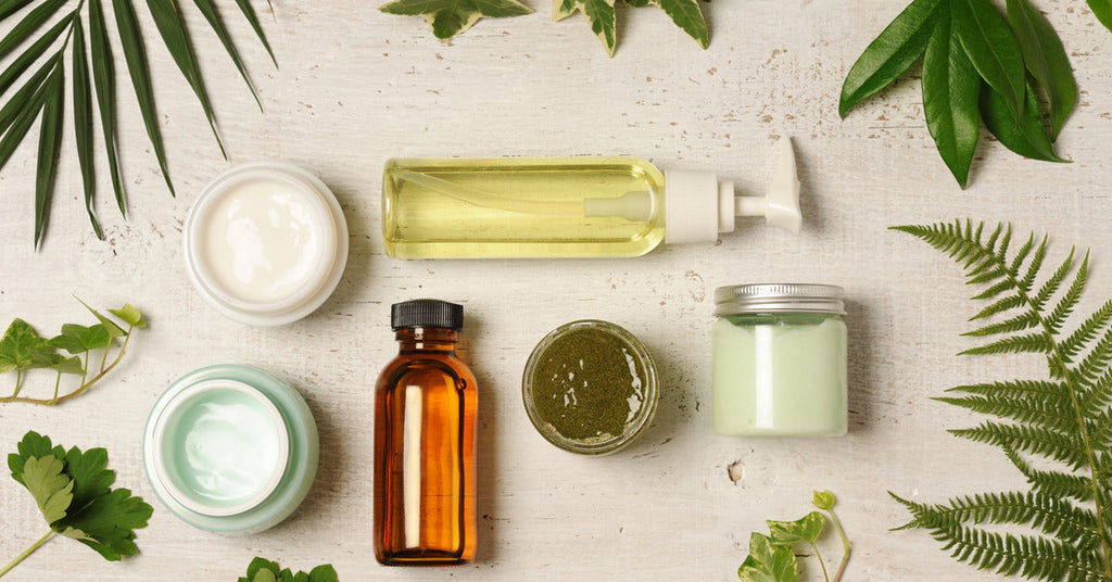 A neat pile of cosmetics that includes two bottles and four containers. They are on a table and surrounded by leaves.