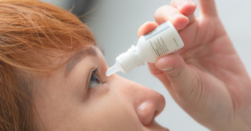 Someone with orange hair is holding a bottle of artificial tears above their right eye as they put a drop in that eye.