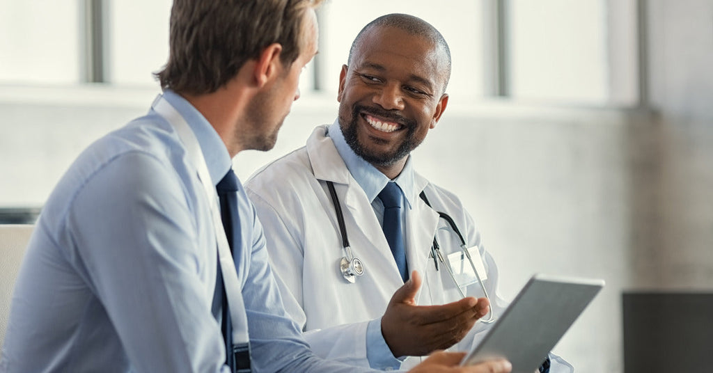 A doctor holding a small tablet while smiling and speaking directly to their patient. They sit in the doctor's office.